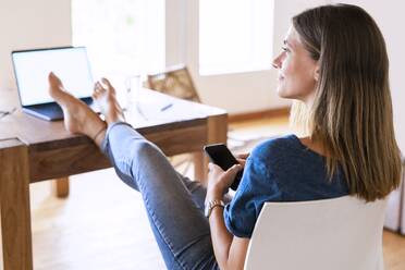 Smiling brunette businesswoman looking away while sitting with smart phone and feet up at table in home office - SBOF02287