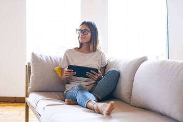 Thoughtful brunette businesswoman sitting with digital tablet on sofa in living room at home - SBOF02300