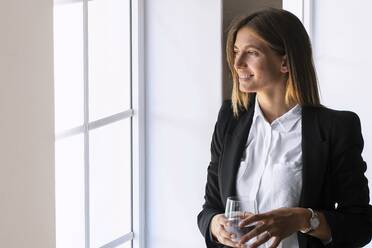 Thoughtful young businesswoman holding drinking glass while looking through window at home - SBOF02310