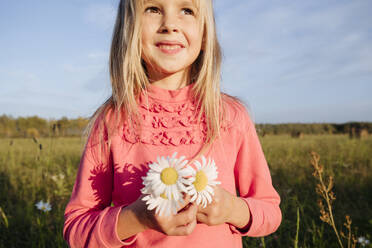 Cute girl holding chamomiles flowers while looking away against sky on sunny day - EYAF01422