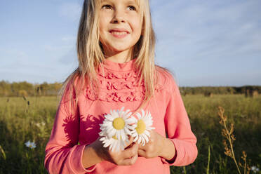 Nettes Mädchen, das Kamillenblüten hält und gegen den Himmel an einem sonnigen Tag wegschaut - EYAF01422