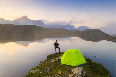 Rear view of man with hat standing outside tent at Obere Schwarziseeli lake at dawn, Furka Pass, Canton Uri, Switzerland, Europe - RHPLF18717