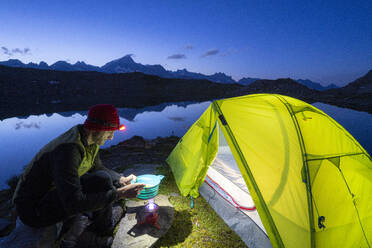Hiker man cooking outside camping tent at Obere Schwarziseeli lake at dusk, Furka Pass, Canton Uri, Switzerland, Europe - RHPLF18723