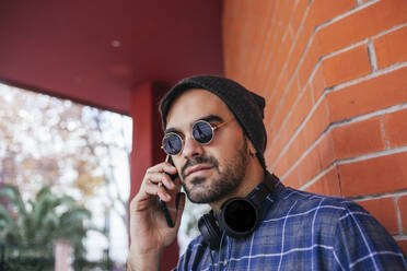 Fashionable young man wearing sunglasses on phone call against brick wall - MGRF00095