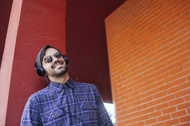 Smiling fashionable young man listening music through headphones against column - MGRF00097