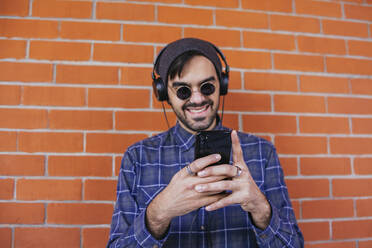 Smiling young man listening music while using smart phone against brick wall - MGRF00106