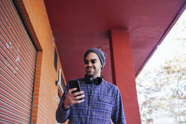 Modischer junger Mann, der lächelt, während er ein Mobiltelefon im Korridor benutzt - MGRF00107