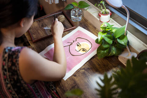 High angle side view of unrecognizable Asian female artist sitting at wooden table and creating picture on paper with paint and brush stock photo