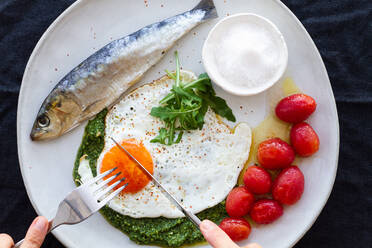 From above crop anonymous person eating fried egg served with salty fish and marinated tomatoes and garnished with pesto sauce - ADSF19491