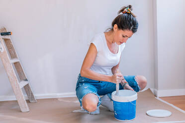 Happy young female taking white paint with brush from bucket while kneeling on floor near wall during renovation at home - ADSF19494