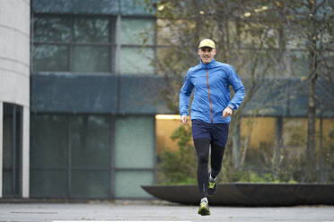 Mature man jogging wearing cap against building - JAHF00074