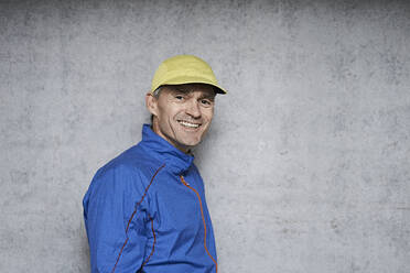 Happy mature man wearing cap standing by gray wall - JAHF00086