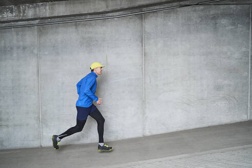 Smiling mature man jogging by gray wall in parking garage - JAHF00088