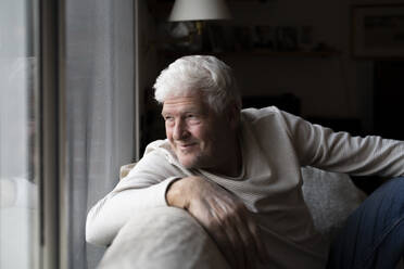 Lächelnder älterer Mann, der durch das Fenster schaut, während er zu Hause auf dem Sofa sitzt - AFVF07878