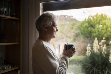 Älterer Mann mit Kaffeetasse, der durch das Fenster schaut, während er zu Hause steht - AFVF07887