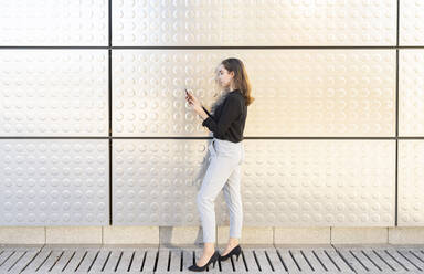 Geschäftsfrau, die ein Mobiltelefon benutzt, während sie an einer silbernen Wand steht - JCCMF00255