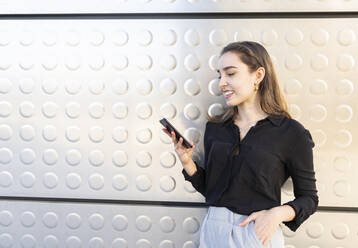 Smiling businesswoman using mobile phone while standing with hands in pockets against silver wall - JCCMF00256