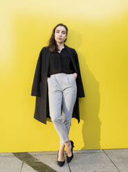 Junge Geschäftsfrau mit Jacke, die mit den Händen in den Taschen vor einer gelben Wand steht - JCCMF00261