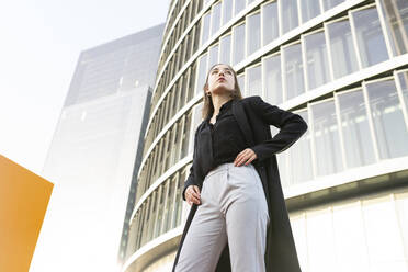 Confident businesswoman looking away while standing with hand on hip in city - JCCMF00267