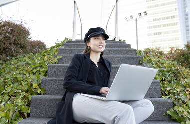 Junge Geschäftsfrau mit Mütze, die wegschaut, während sie mit einem Laptop auf einer Treppe sitzt - JCCMF00271