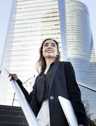 Lächelnde junge Geschäftsfrau mit Laptop auf einer Treppe in der Stadt stehend - JCCMF00273