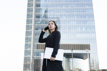 Young businesswoman with laptop talking on mobile phone while standing in city - JCCMF00278