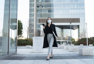 Businesswoman wearing face mask using mobile phone while walking on footpath - JCCMF00283