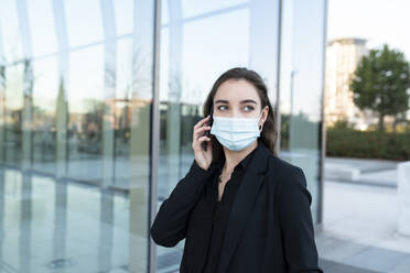 Young businesswoman wearing protective face mask talking on mobile phone while standing against building - JCCMF00285