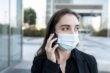 Businesswoman with protective face mask looking away while talking on mobile phone outdoors - JCCMF00287