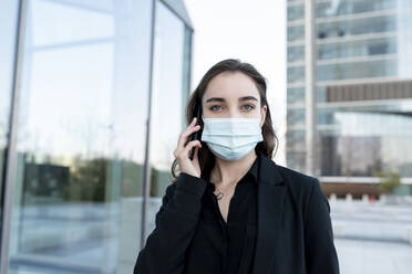 Young businesswoman with protective face mask staring while talking on mobile phone standing outdoors - JCCMF00288