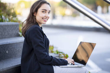 Lächelnde Geschäftsfrau, die einen Laptop benutzt und auf einer Treppe sitzt - JCCMF00304