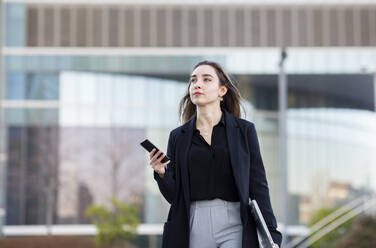 Young businesswoman with laptop using mobile phone while walking outdoors - JCCMF00309