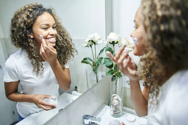 Schöne lächelnde Frau trägt Gesichtscreme auf und betrachtet ihr Spiegelbild im Badezimmer - KIJF03503