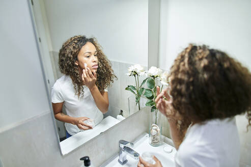 Schöne Frau trägt Gesichtscreme auf und betrachtet ihr Spiegelbild im Badezimmer - KIJF03504