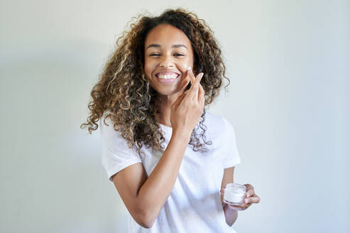 Cheerful young woman applying facial cream against white wall - KIJF03505