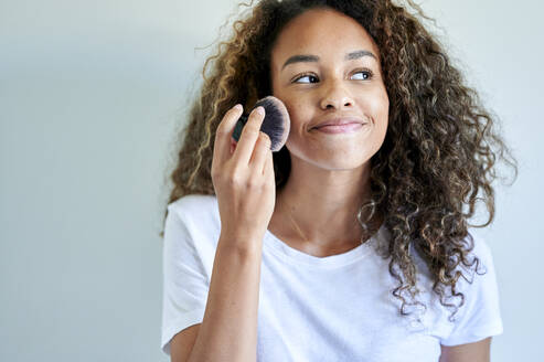 Smiling young woman applying face powder with make-up brush against white wall - KIJF03507
