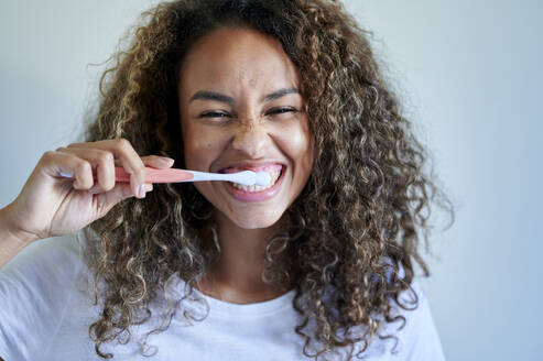 Cheerful young woman enjoying while brushing teeth against wall - KIJF03510