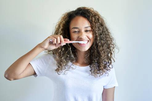 Happy young woman enjoying while brushing teeth against wall - KIJF03511