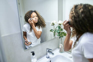 Junge Frau trägt Gesichtspuder mit einem Make-up-Pinsel auf, während sie sich im Badezimmer im Spiegel betrachtet - KIJF03512