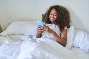 Happy young woman using smart phone while relaxing on bed in bedroom at home - KIJF03516