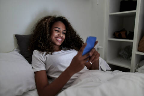 Smiling young woman using smart phone while relaxing on bed in bedroom at home stock photo