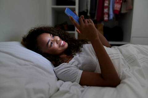 Happy young woman using smart phone while lying on bed in bedroom at home stock photo
