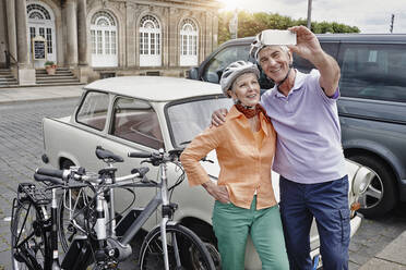 Lächelndes Paar mit Fahrradhelm, das ein Selfie mit einem Trabant macht, während es in Dresden steht, Deutschland - RORF02564