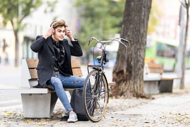 Junger Mann stellt drahtlose Kopfhörer ein, während er auf einer Bank mit dem Fahrrad in der Stadt im Herbst sitzt - GGGF00529