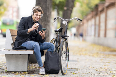 Handsome young man using smart phone while drinking coffee by bicycle in city during autumn - GGGF00530