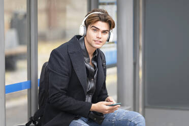 Hübscher junger Mann hört Musik, während er an der Bushaltestelle sitzt - GGGF00531