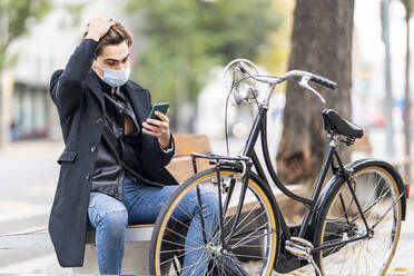 Young man with hand in hair using smart phone while sitting on bench by bicycle in city during COVID-19 - GGGF00532