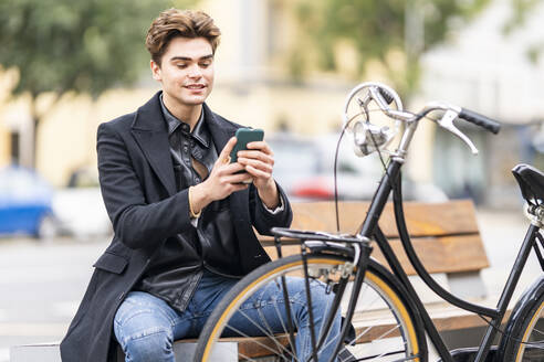 Junger Mann, der auf einer Bank in der Nähe eines Fahrrads in der Stadt sitzt und mit seinem Mobiltelefon SMS schreibt - GGGF00535