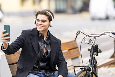 Smiling handsome young man taking selfie through smart phone while sitting by bicycle in city - GGGF00536