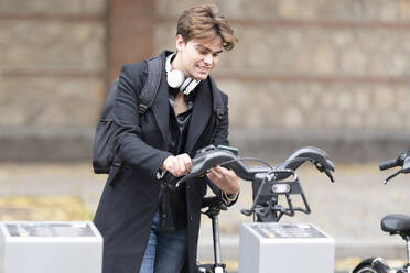 Smiling young handsome man using smart phone for unlocking electric sharing bicycle in city - GGGF00541